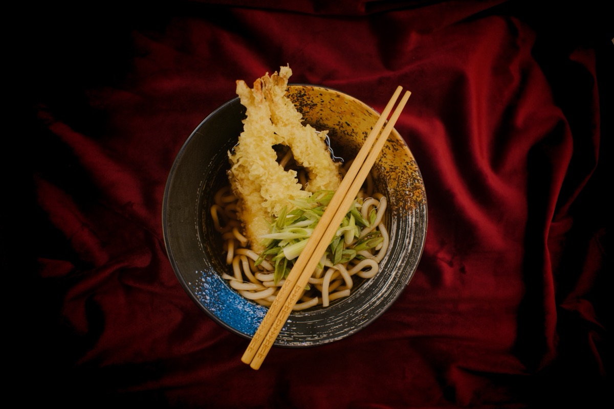 Tempura on udon in a black bowl with chopsticks, on red velvet